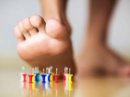 Photograph of a human foot hovering above several push pin needles laying upright on a floor
