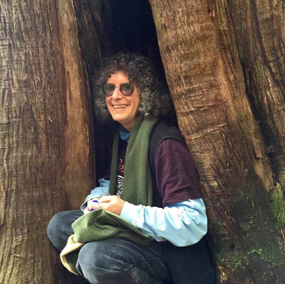 Photograph of Laurie smiling, crouching down in between a crevice of a trees' buttress